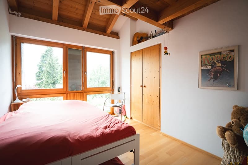 Cozy bedroom with wooden ceiling beams, large windows, a bed with pink bedding, a wooden wardrobe, a guitar on a shelf, a small table with a lamp, and a wicker basket with toys.