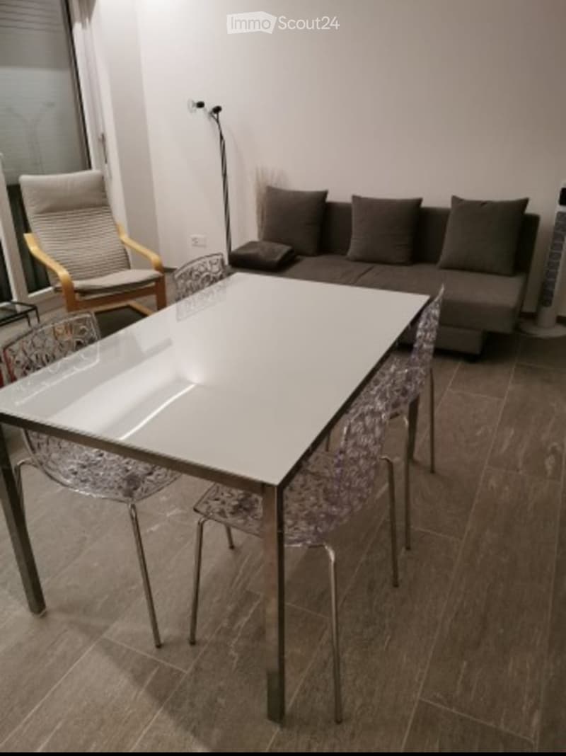 Modern living room with a sofa, glass table, chairs, and a floor lamp near a large window.