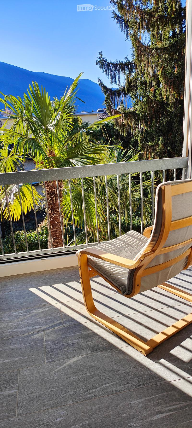 Balcony with a wooden chair, metal railing, and lush greenery in the background, overlooking a scenic mountain view.