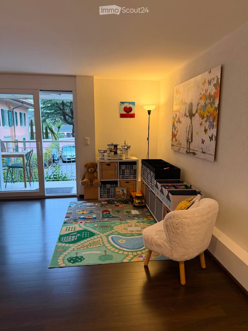 A cozy, well-lit room with wooden flooring, a large play mat, a plush chair, and various toys and storage units.