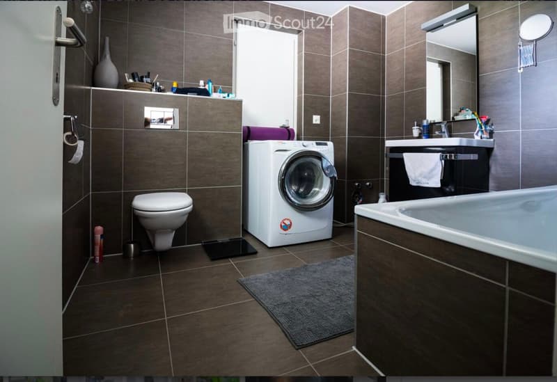 Modern bathroom with dark brown tiles, a toilet, a washing machine, a bathtub, a sink with a mirror, and a towel on a rack.