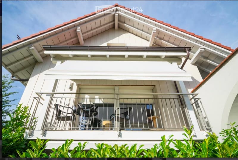 Balcony with metal railing, featuring a table and chairs, surrounded by greenery.