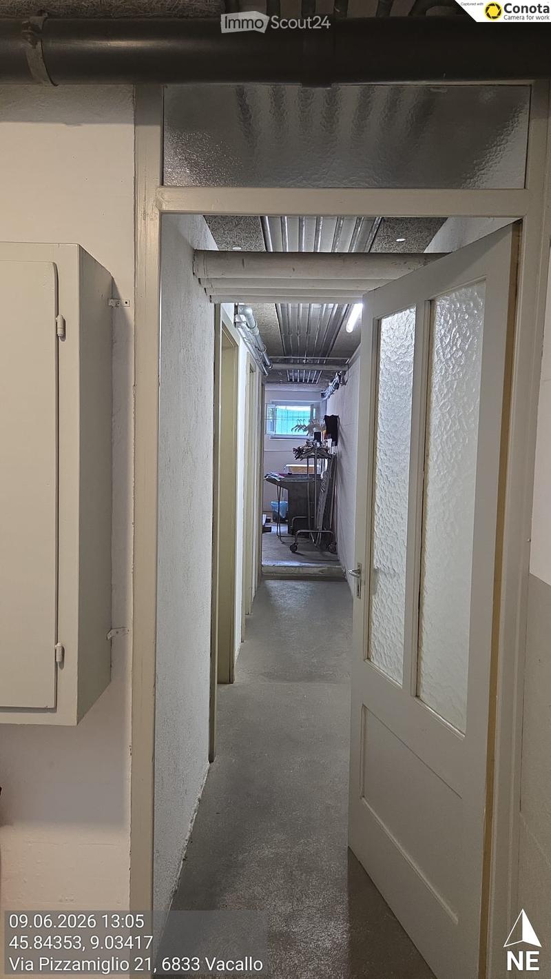 A long, narrow hallway with white walls, a gray carpeted floor, and a ceiling with exposed pipes and lighting. The hallway has several doors on both sides and leads to a room with a window and some furniture visible at the end.