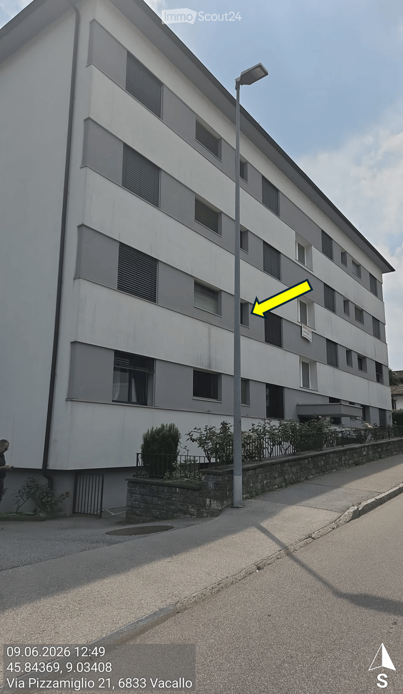Multi-story residential building with grey and white facade, multiple windows, and a small garden area at the base.
