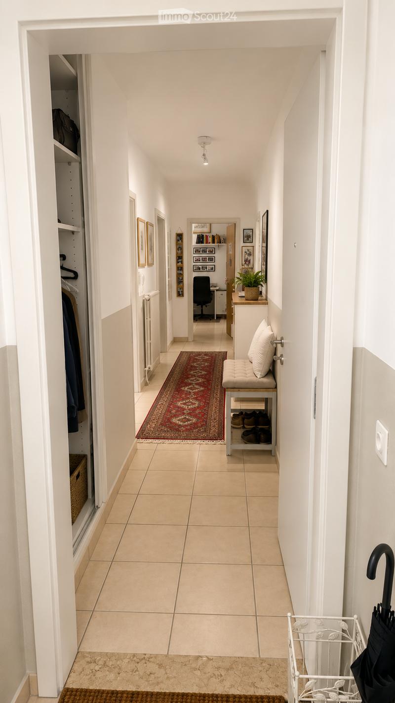 A well-lit hallway with tiled flooring, white walls, and a small bench with shoes underneath. A red patterned rug is placed on the floor, and a small umbrella stand is visible on the right side.