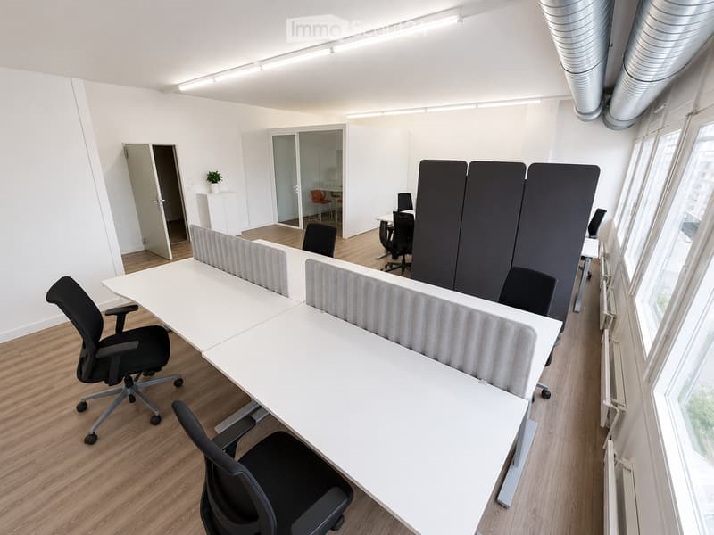 Modern office space with white tables, black chairs, and privacy screens. Large windows provide natural light, and the floor is wooden.