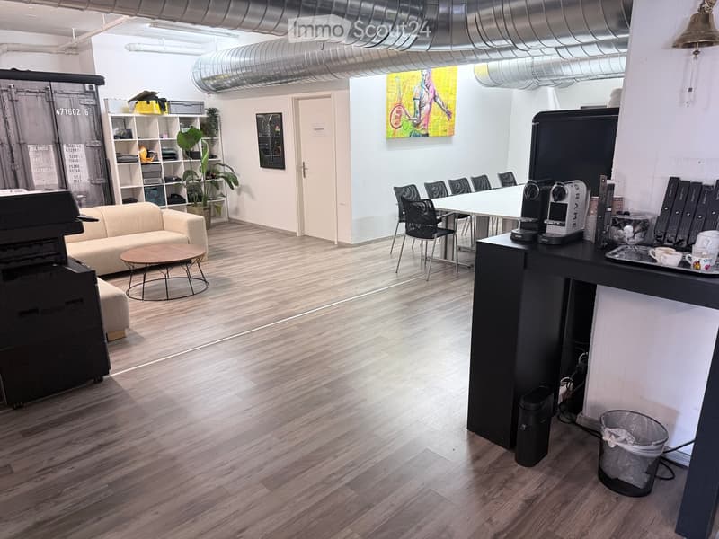 Modern office space with a sofa, coffee table, chairs, a large table, printer, and a small kitchen area with a coffee machine and cups.