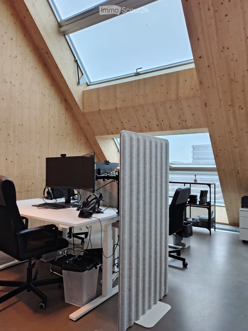 Modern office space with wooden walls, large skylight windows, and a desk setup with dual monitors, keyboard, mouse, and chairs on both sides of a screen divider.