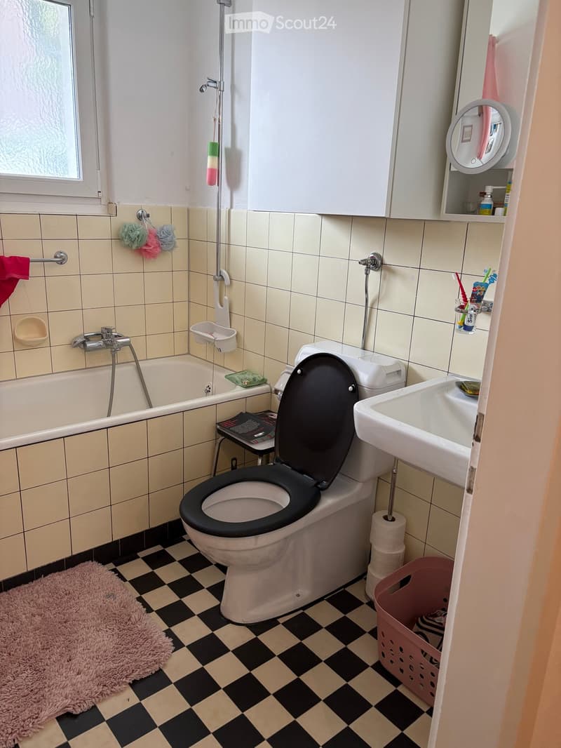 Bathroom with beige tiled walls, black and beige checkered floor, toilet, sink, bathtub, mirror, and various accessories including a bathrobe, bath mat, and toothbrush holder.