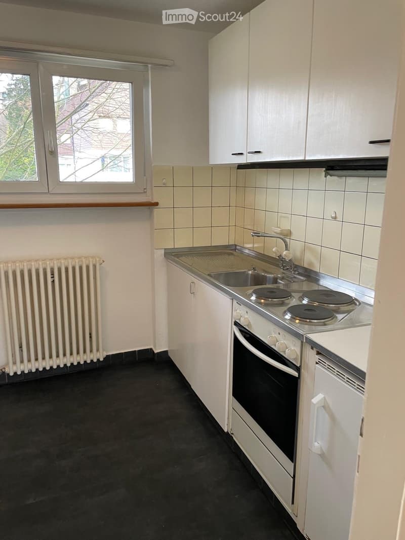 Compact kitchen with white cabinets, a sink, a four-burner electric stove, and an oven. The room features tiled walls, a window, and a radiator.