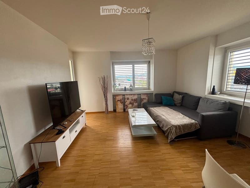 Modern living room with wooden flooring, a grey sofa with a throw, a white coffee table, a flat-screen TV on a white stand, and a hanging chandelier.