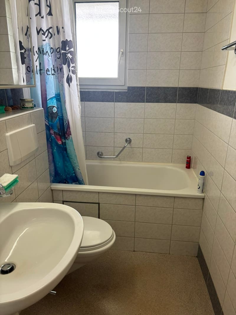 Bathroom with a bathtub, toilet, sink, and a shower curtain. The walls are tiled with a mix of light and dark tiles, and there is a window above the bathtub.