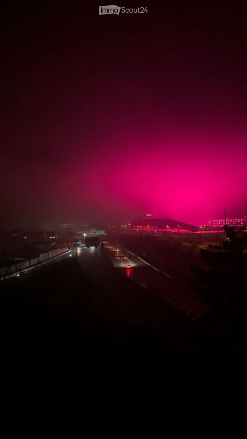 Nighttime view of a highway with traffic, distant illuminated building, and pink sky.