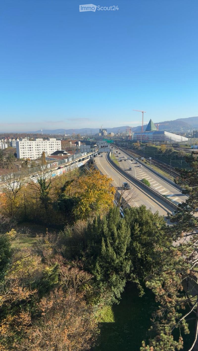 A panoramic view of a cityscape with a multi-lane highway, railway tracks, and surrounding buildings under a clear blue sky.