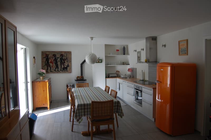 Bright kitchen with white cabinets, an orange refrigerator, a dining table with chairs, and a pendant light. Features a wooden sideboard, a painting, and a vase on a shelf.