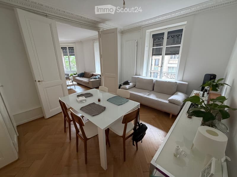 Bright, open-plan living and dining area with wooden flooring, white walls, large windows, and a balcony view. Features a white dining table with four chairs, a grey sofa, and a small plant on a side table.