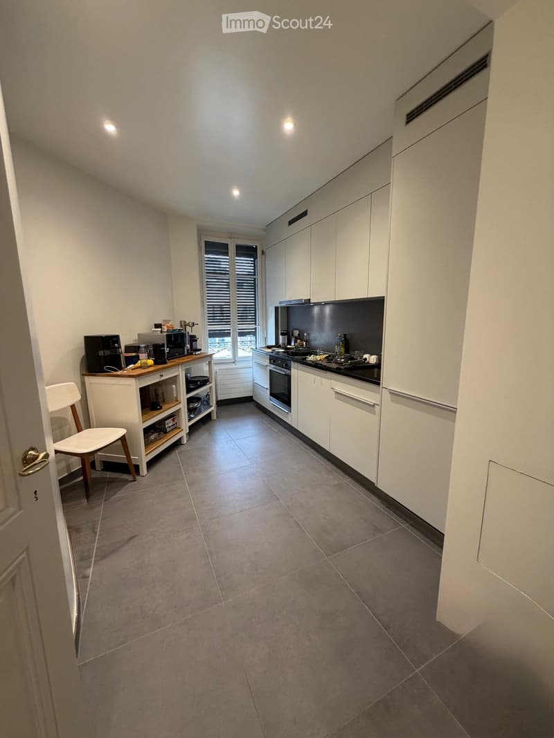 Modern kitchen with white cabinets, grey tiled floor, and various appliances including a coffee maker, oven, and sink. A small wooden chair and countertop are also visible.