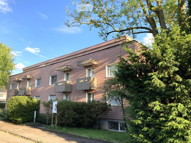 Two-story building with pink siding, multiple windows, and balconies, surrounded by trees and shrubs.