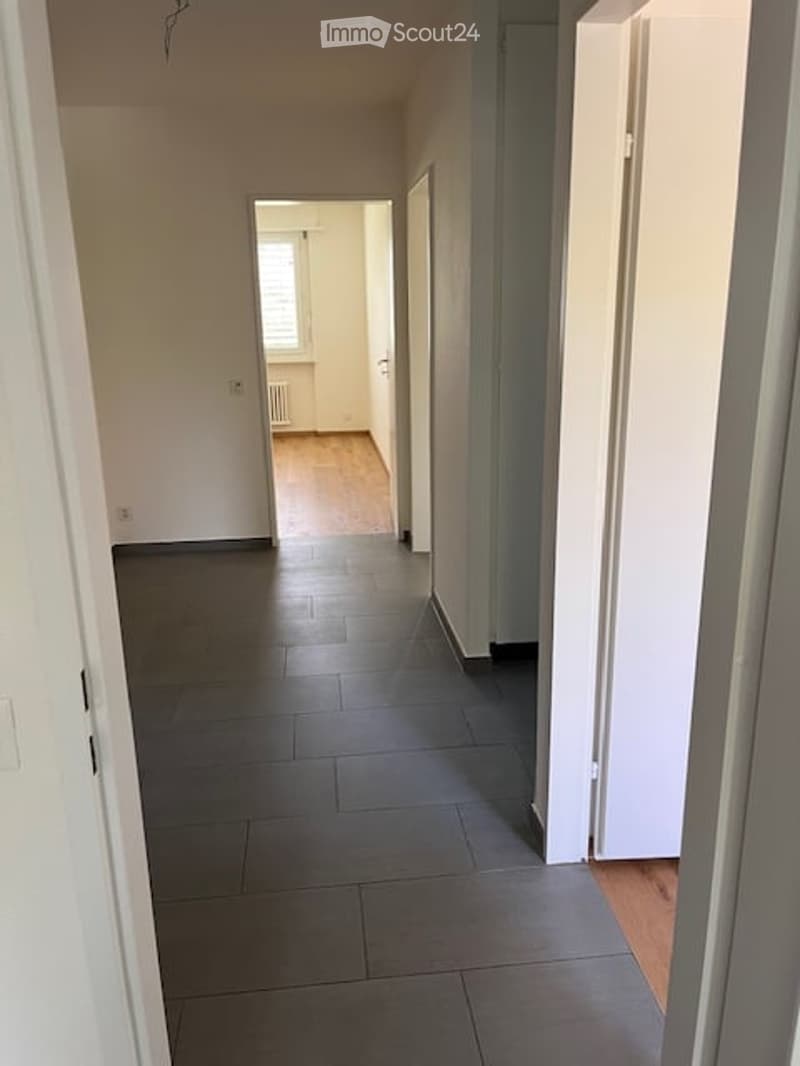 Empty hallway with grey tiled floor, white walls, and a visible room at the end with wooden flooring and a window.