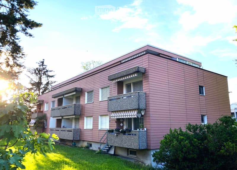 Two-story pink building with multiple balconies, white shutters, and a small garden area in front.