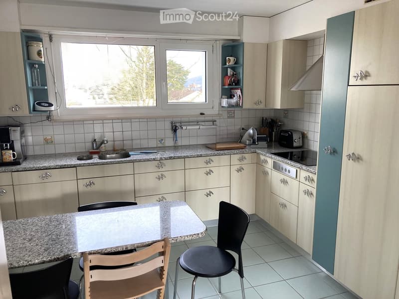 Bright kitchen with cream-colored cabinets, granite countertops, and a small dining area with chairs and a table. Appliances include a coffee maker, toaster, and oven. Windows provide natural light.