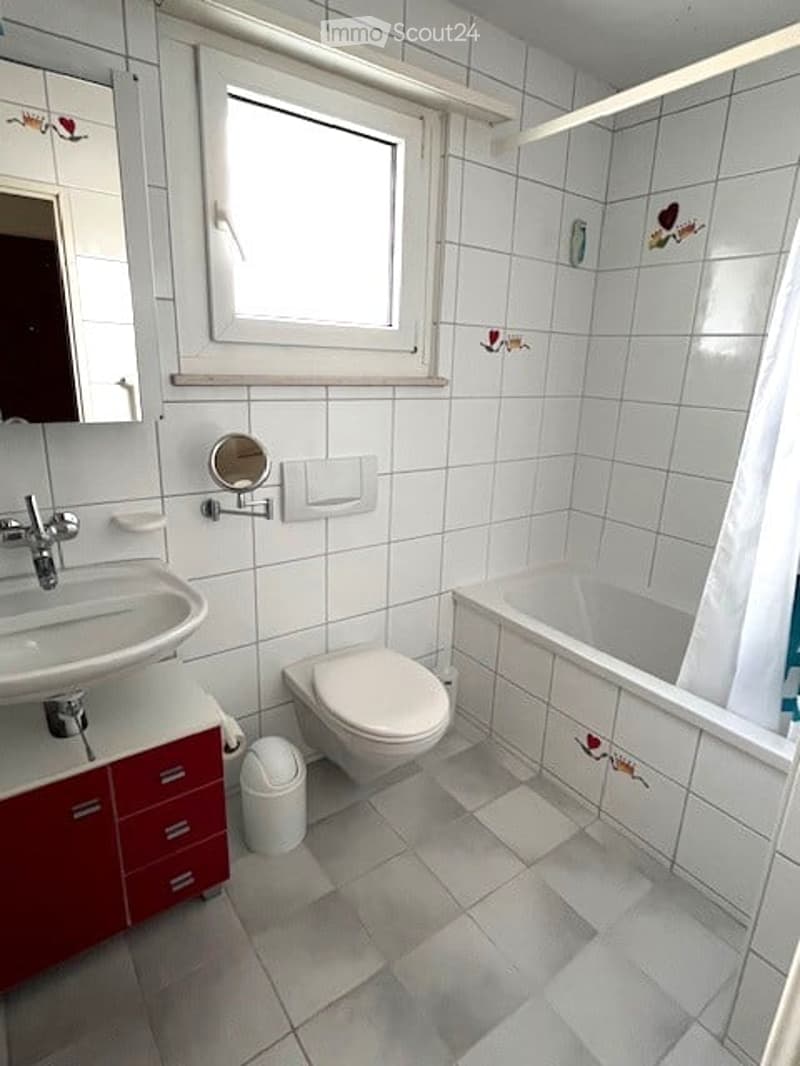Bright bathroom with white tiles, a sink, toilet, bathtub, and a window above the bathtub. Red cabinetry is visible under the sink.