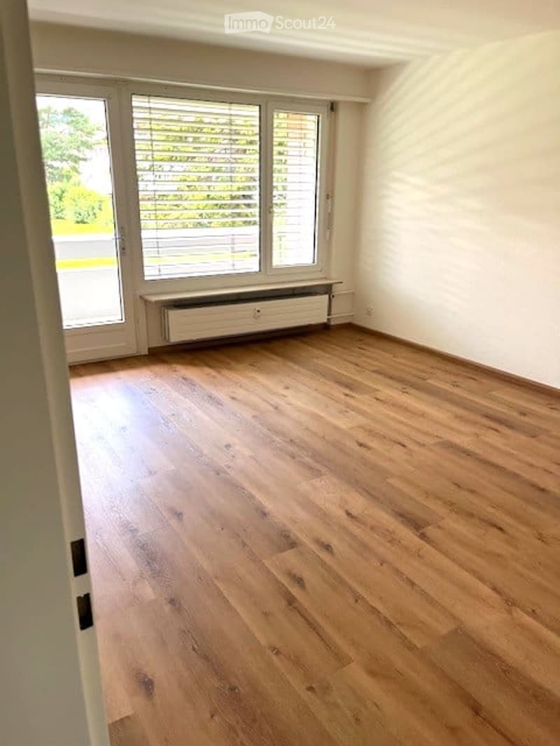 Empty room with wooden flooring, large windows with blinds, and a radiator under the windows.