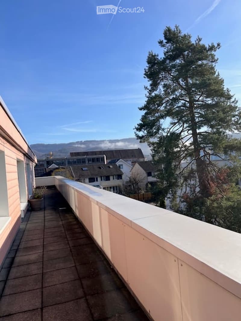 Balcony with white railing, tiled floor, overlooking a residential area with trees and distant mountains.