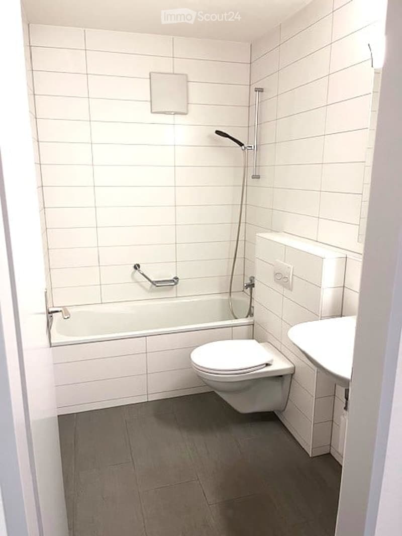 Modern bathroom with white tiled walls and floor, featuring a bathtub, toilet, sink, and a showerhead with a handrail.