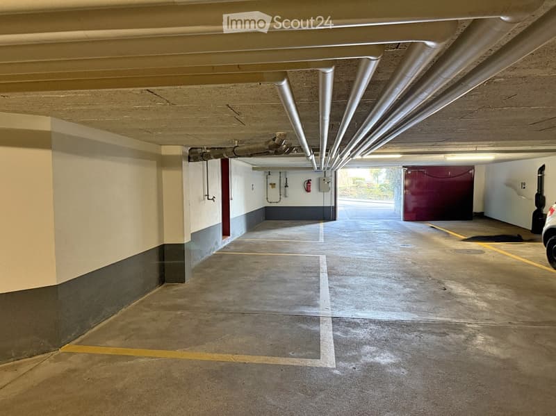 Underground parking garage with marked parking spaces, concrete floors, and exposed pipes. A red storage unit is visible in the background.