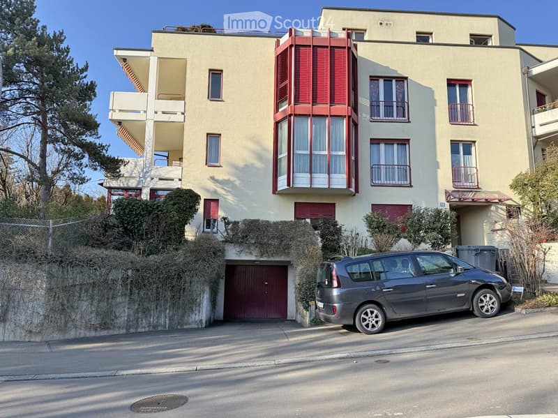 Four-story residential building with beige facade, red shutters, balconies, and a garage entrance. A parked car is visible in front of the building.