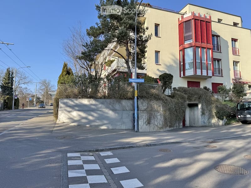 Multi-story residential building with red shutters and balconies, located on a quiet street with trees and a sidewalk.