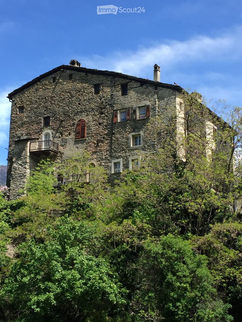 A rustic stone house with multiple windows, shutters, and a small balcony, surrounded by lush greenery.
