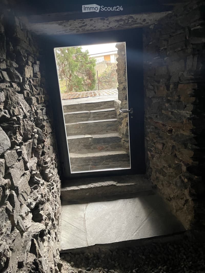 A narrow stone-walled corridor with a large window opening to an outdoor staircase and garden area.