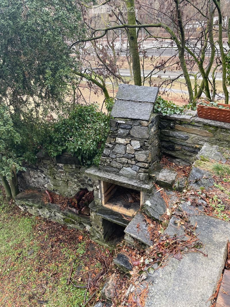Stone outdoor fireplace with a raised platform, surrounded by trees and greenery, with a paved area and a small wooden storage space underneath.
