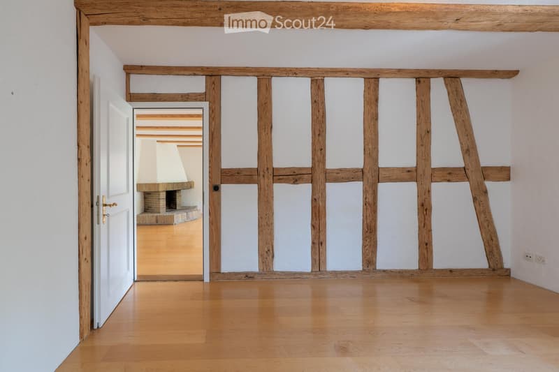 Empty room with wooden floor, white walls, exposed wooden beams, and an open doorway leading to another room with a fireplace.
