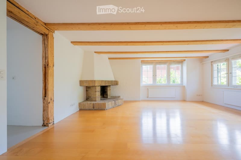 Empty room with wooden floor, exposed wooden beams, and a brick fireplace with a white chimney. Large windows provide natural light.