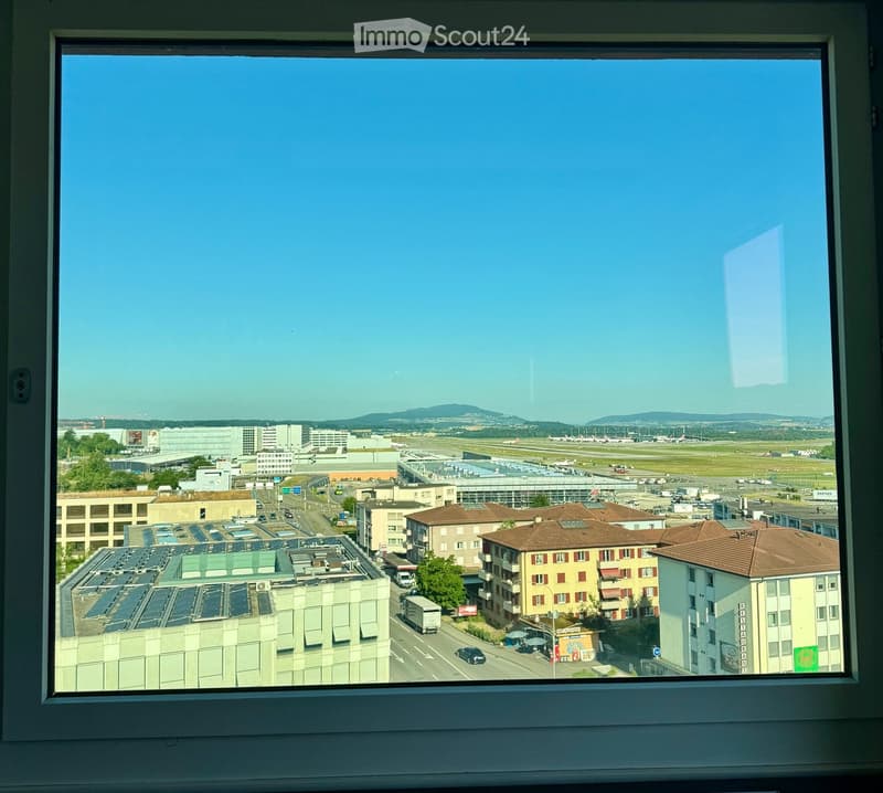View from a window showing an airport with runways, buildings, and distant mountains under a clear blue sky.