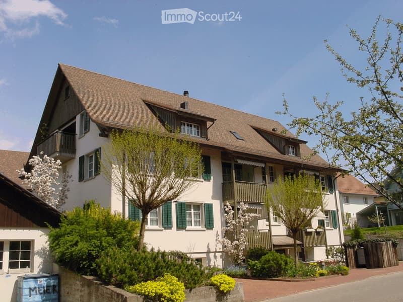 A two-story house with a brown tiled roof, white walls, green shutters, and multiple windows. The house features balconies on the upper floor and is surrounded by well-maintained gardens and trees.