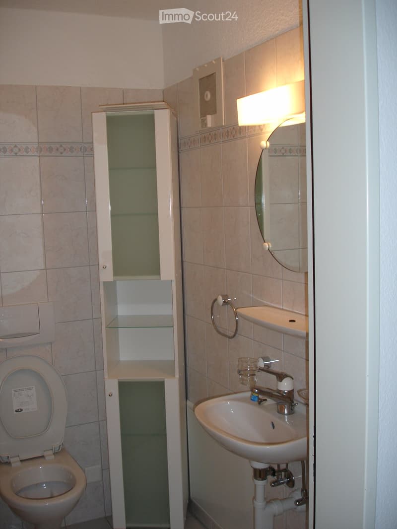 A small bathroom with a toilet, sink, and a mirror. The bathroom has white tiles on the walls and a white cabinet with glass doors.