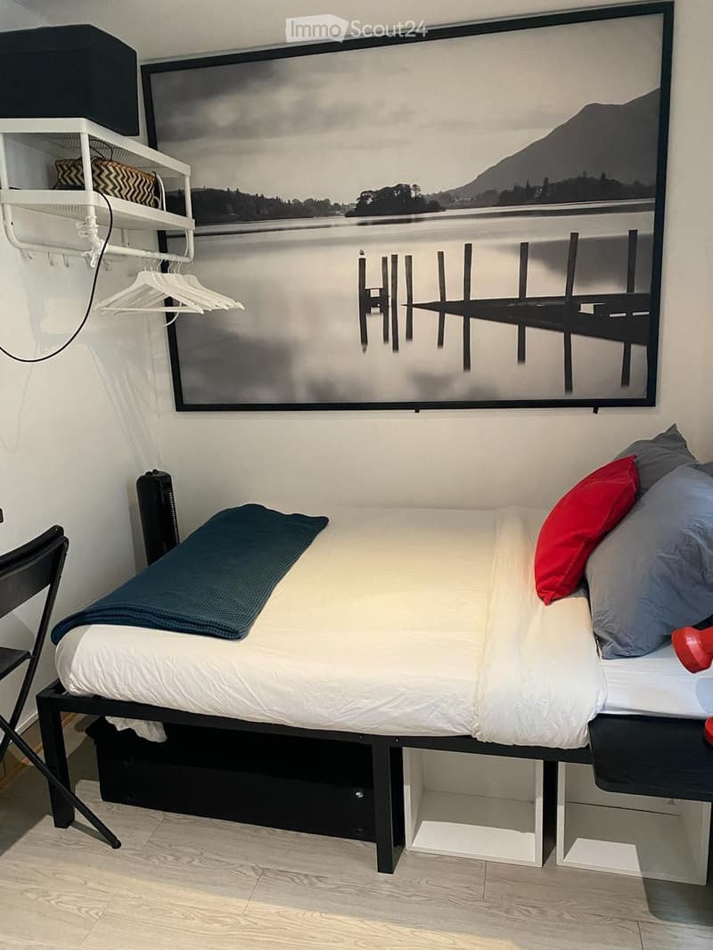 A modern, compact bedroom with a single bed, a nightstand, a chair, and a large picture frame above the bed. The bed is neatly made with white sheets and red and blue pillows.