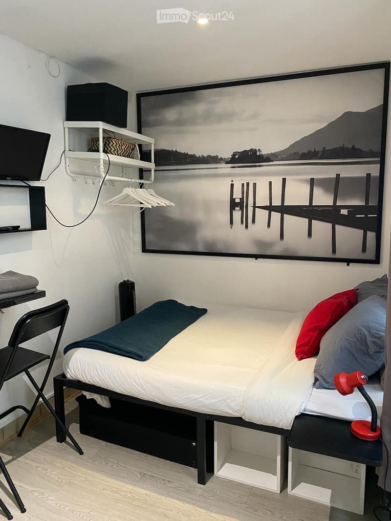 A compact bedroom with a single bed, nightstand, lamp, chair, table, shelves, and a large wall-mounted television.