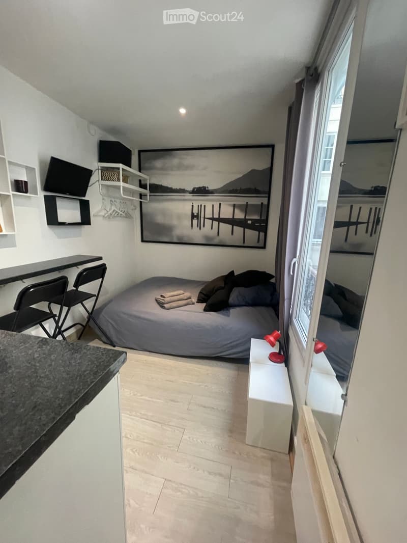 A compact, modern bedroom with a bed, a small table, chairs, shelves, a lamp, and a screen. The room features light wooden flooring and white walls.