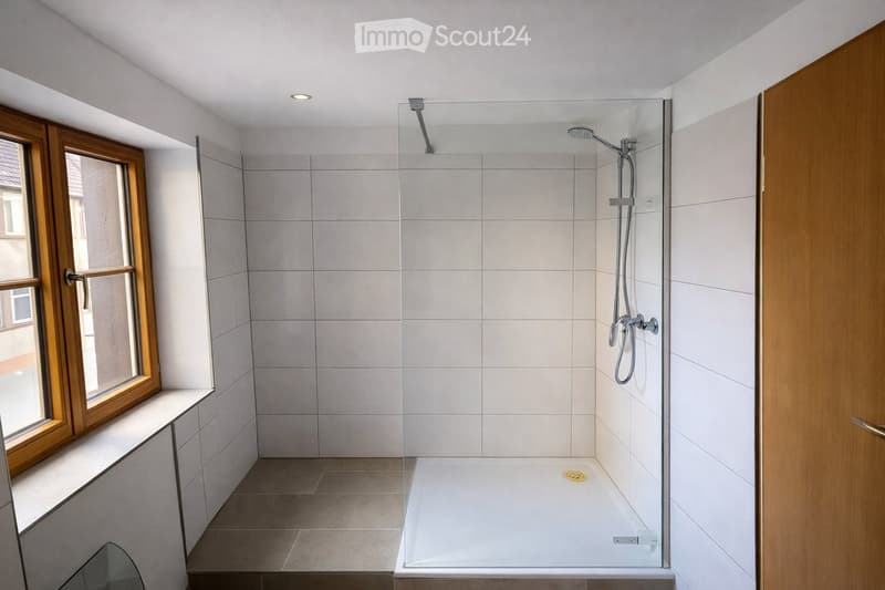 Modern bathroom with a bathtub, glass-enclosed shower, and a window with wooden frame. The walls and floor are tiled in neutral tones.