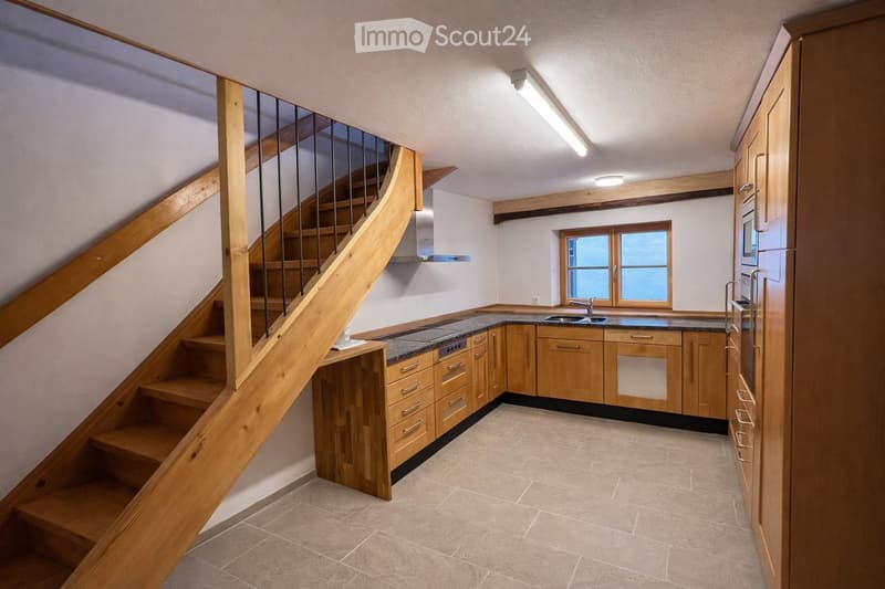 Modern kitchen with wooden cabinets, granite countertops, and a built-in sink. A wooden staircase with a glass railing leads to an upper level.