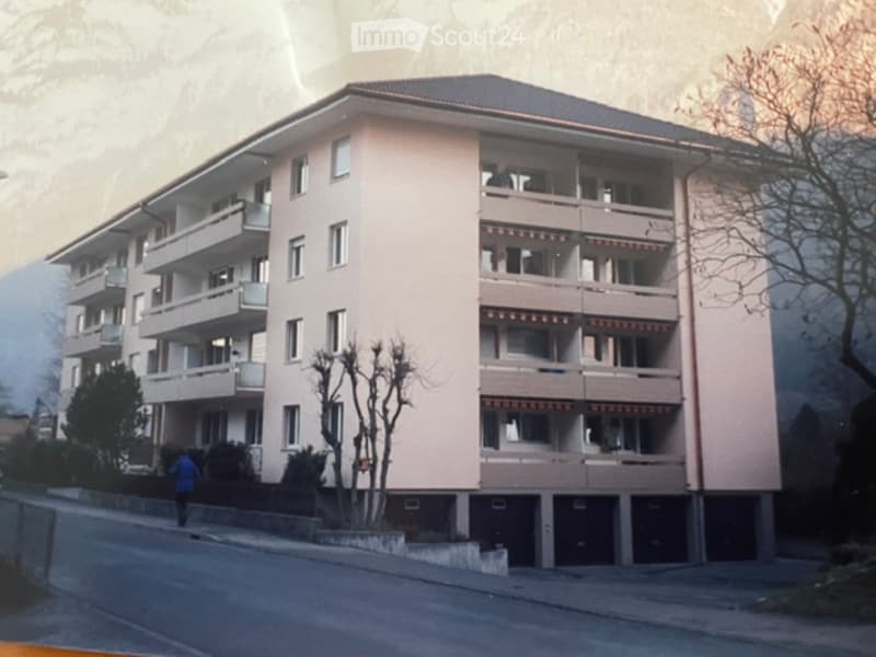 Multi-story residential building with multiple windows and balconies, surrounded by trees and a road.