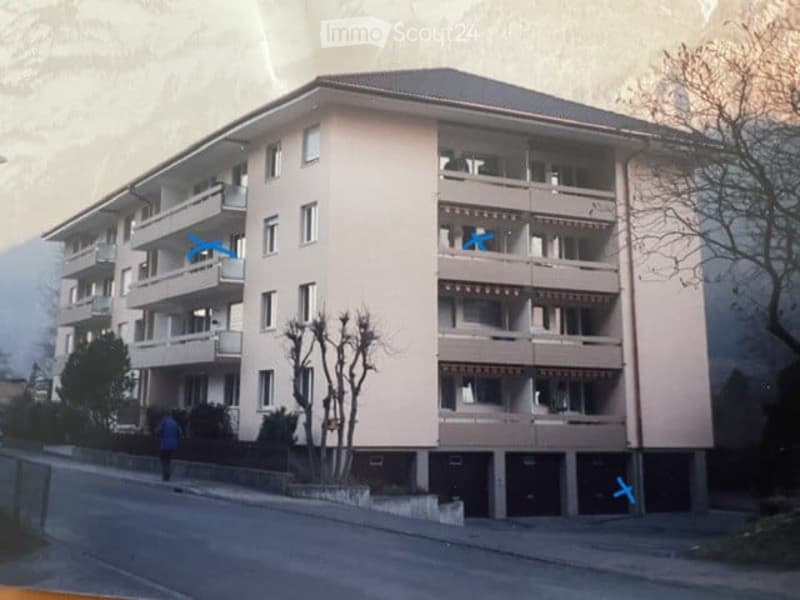 Multi-story residential building with multiple balconies and windows, surrounded by trees and a road.