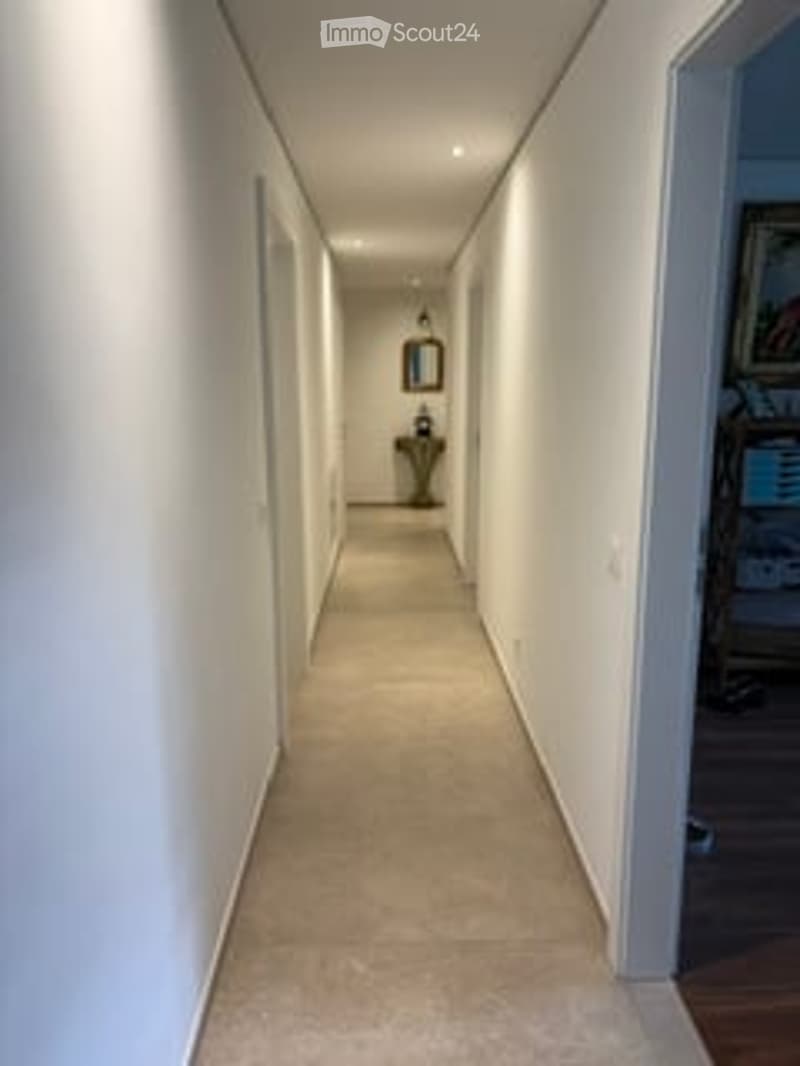 A long, narrow hallway with white walls, a beige tiled floor, and recessed lighting. A small decorative item is placed on the floor at the end of the hallway.