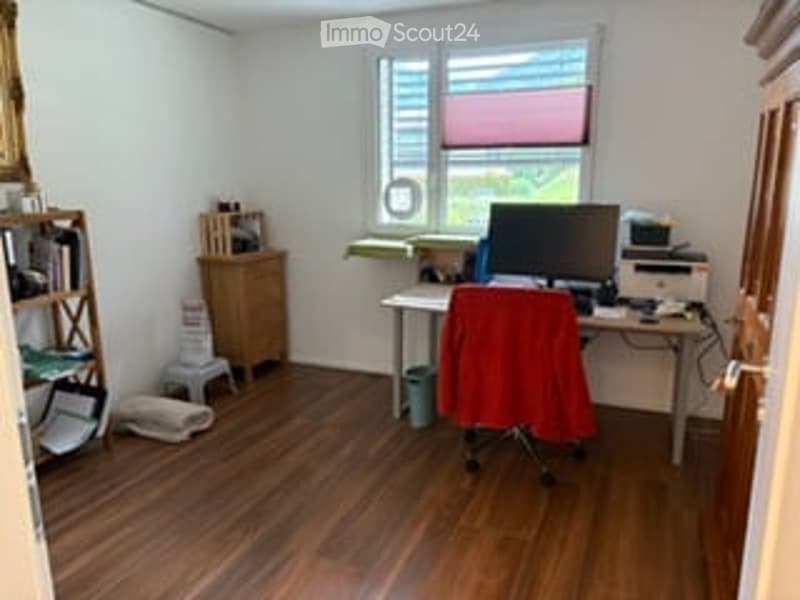 A small, well-lit room with wooden flooring, a desk with a computer monitor, a red chair, a printer, a bookshelf, and a small cabinet.