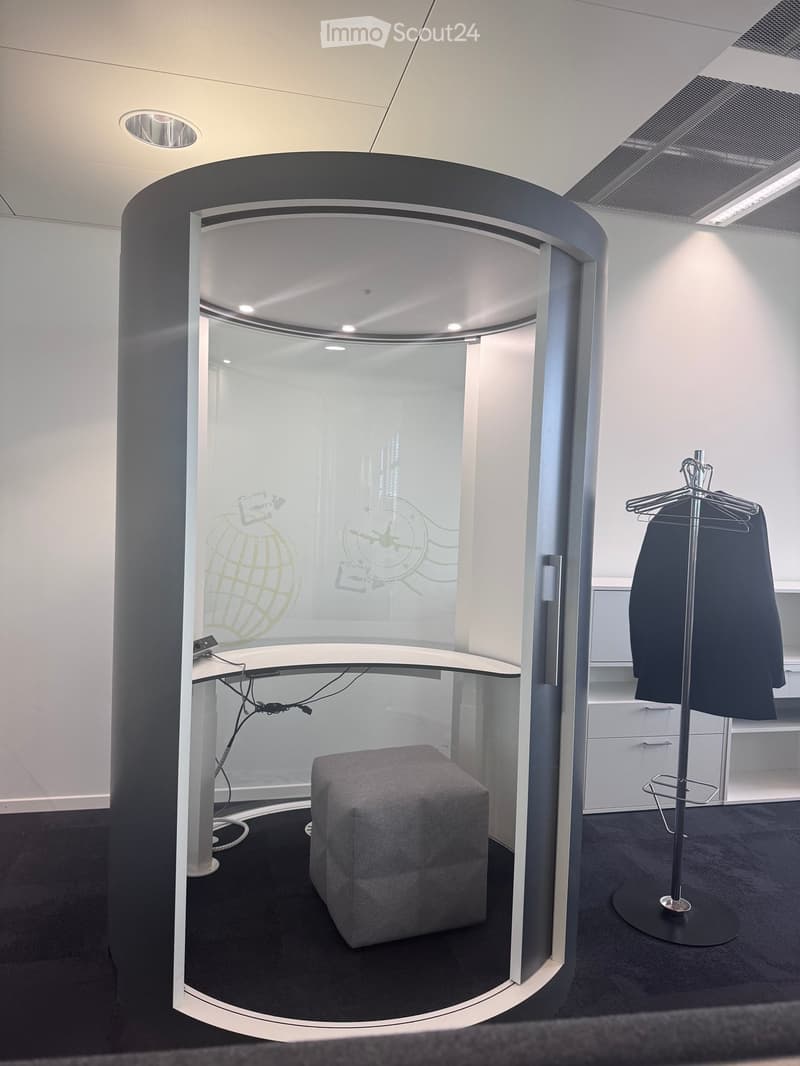 A modern, enclosed office space with a round, glass-walled booth. Inside, there is a grey stool, a sleek white table with cables, and a coat rack with a dark jacket hanging on it. The booth features a contemporary design with a black and white color schem